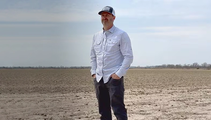 A man wearing a light striped shirt, dark pants, and a black-and-white baseball cap is looking into the distance on a dry field.