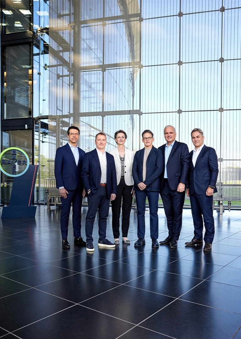 The board of Bayer, consisting of five men and one woman, is posing in business attire for a group photo inside a modern building in front of a glass wall, with a lawn visible behind it. The Bayer Group logo is visible on the left side.