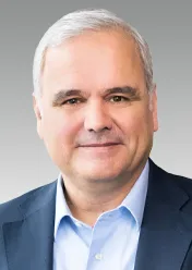 Stefan Oelrich is wearing a white shirt, a black blazer, and is smiling at the camera.