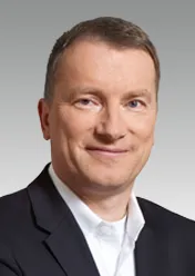 Wolfgang Nickl is wearing a white shirt, a black blazer, and is smiling at the camera.