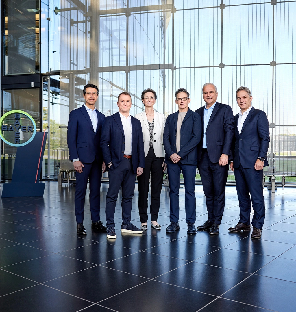 Der Vorstand von Bayer, bestehend aus fünf Männern und einer Frau, posiert in Businesskleidung für ein Gruppenfoto in einem modernen Gebäude vor einer Glaswand, hinter der eine Wiese zu sehen ist. Auf der linken Seite ist das Logo der Bayer-Gruppe sichtbar.