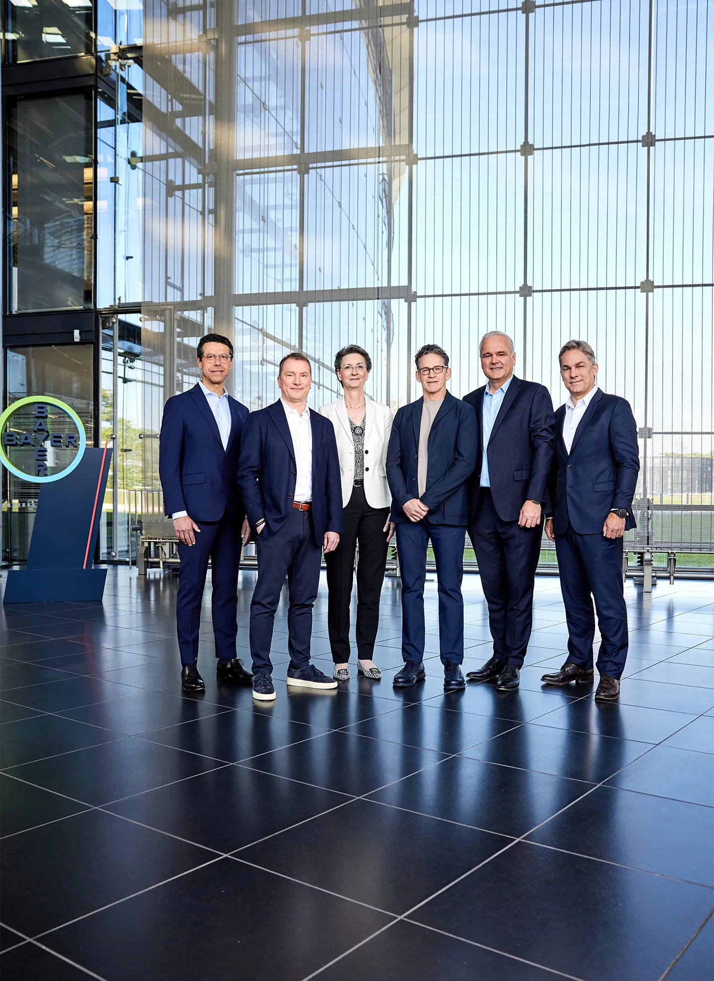 Der Vorstand von Bayer, bestehend aus fünf Männern und einer Frau, posiert in Businesskleidung für ein Gruppenfoto in einem modernen Gebäude vor einer Glaswand, hinter der eine Wiese zu sehen ist. Auf der linken Seite ist das Logo der Bayer-Gruppe sichtbar.