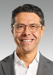 Julio Triana trägt ein weißes Hemd, einen grauen Sakko und eine Brille und lächelt in die Kamera.